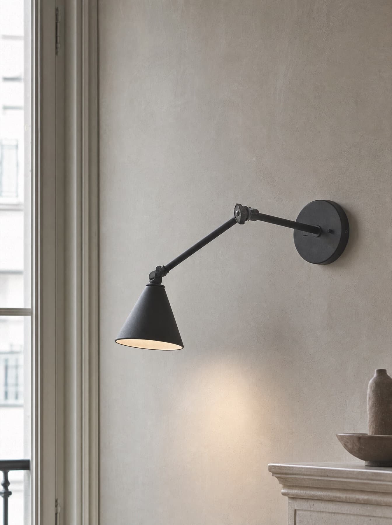 Applique Sète à bras articulé en acier noir mat montée sur un mur à la chaux, flaque de lumière chaude sur l’enduit