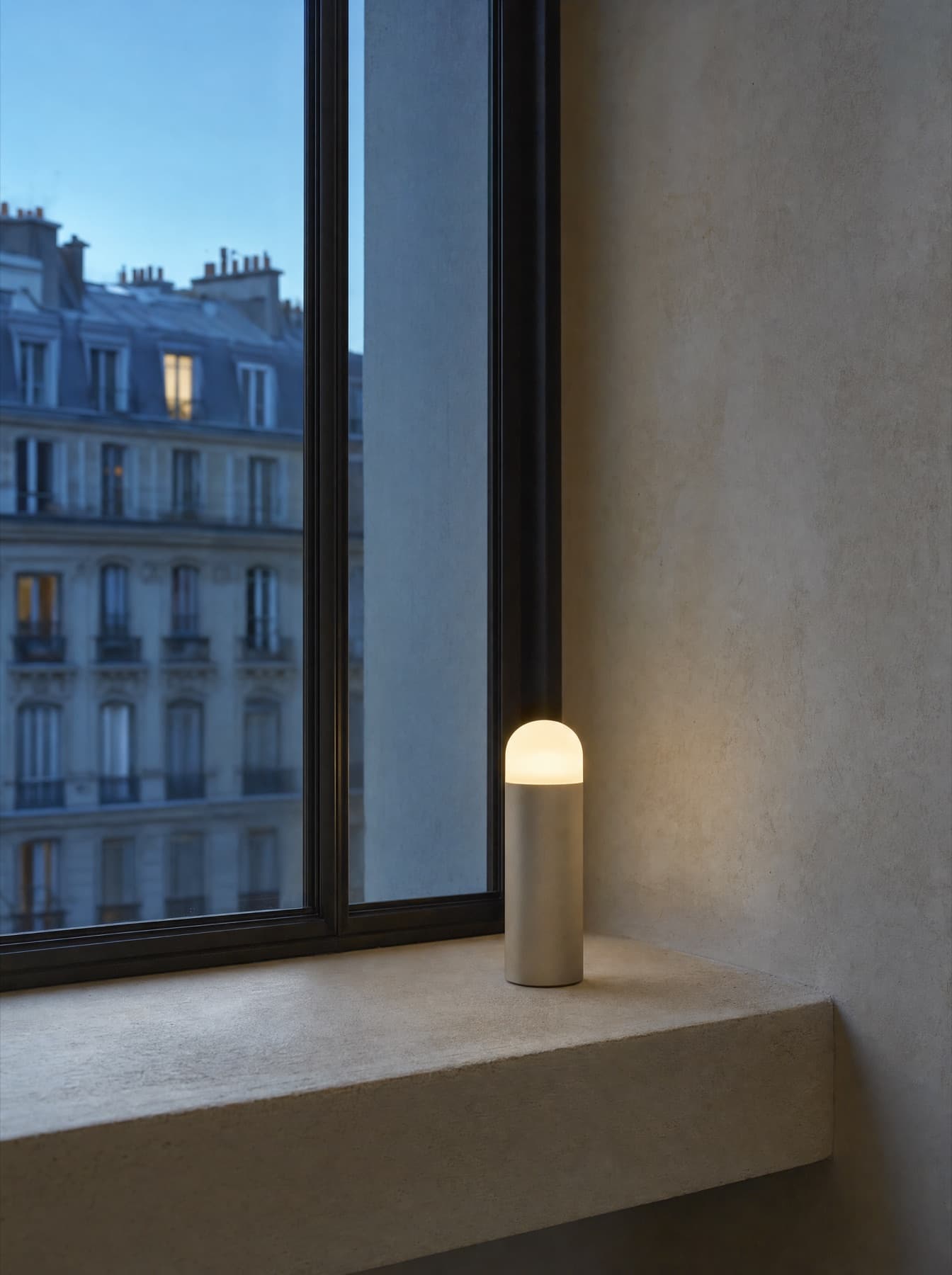 Baladeuse Cassis en aluminium anodisé sable posée sur l’appui d’une fenêtre, dôme diffusant allumé au crépuscule