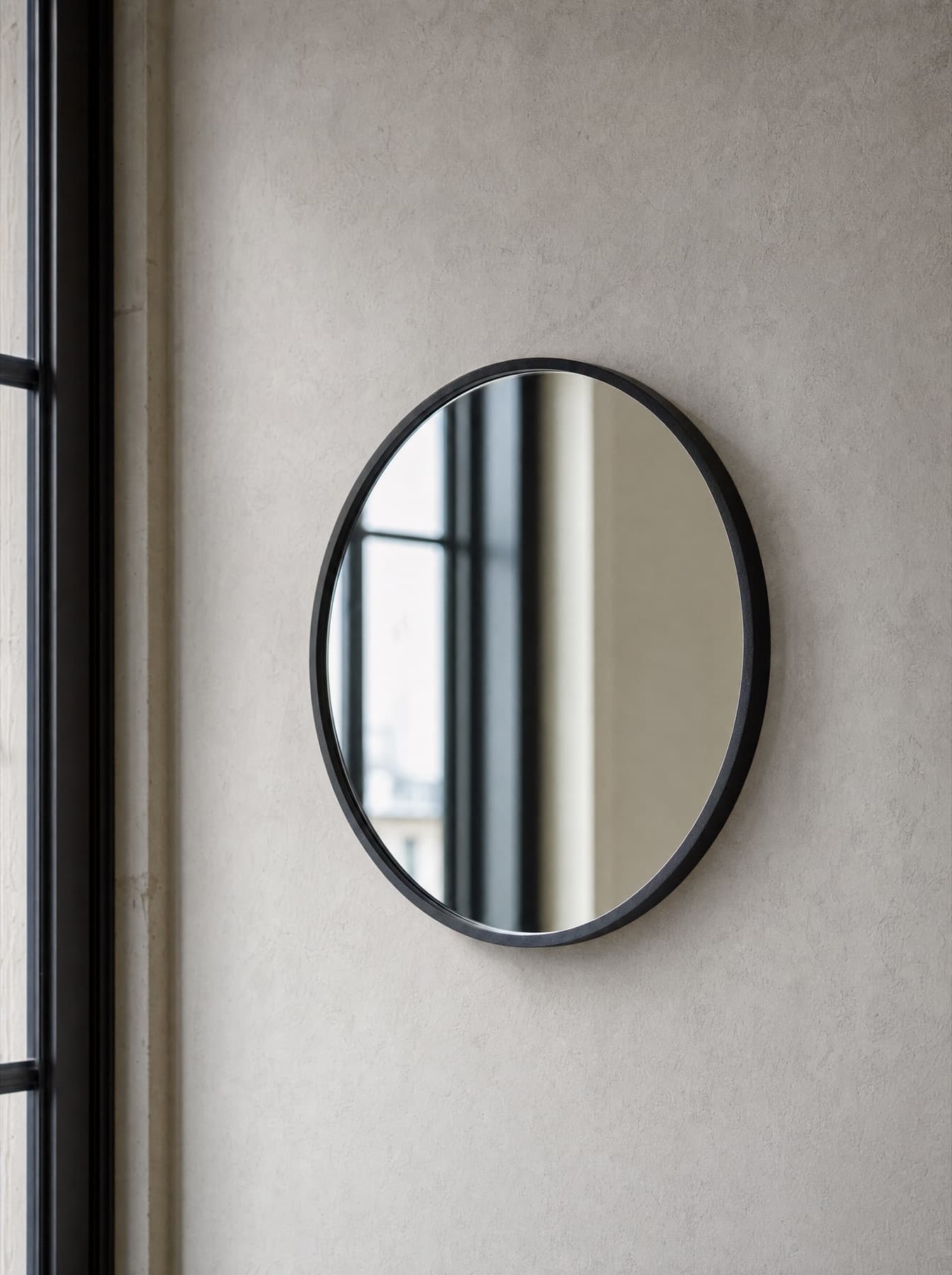Miroir Bréhat accroché sur un mur à la chaux, cadre rond fin en acier laqué noir mat, reflet flou d’une fenêtre