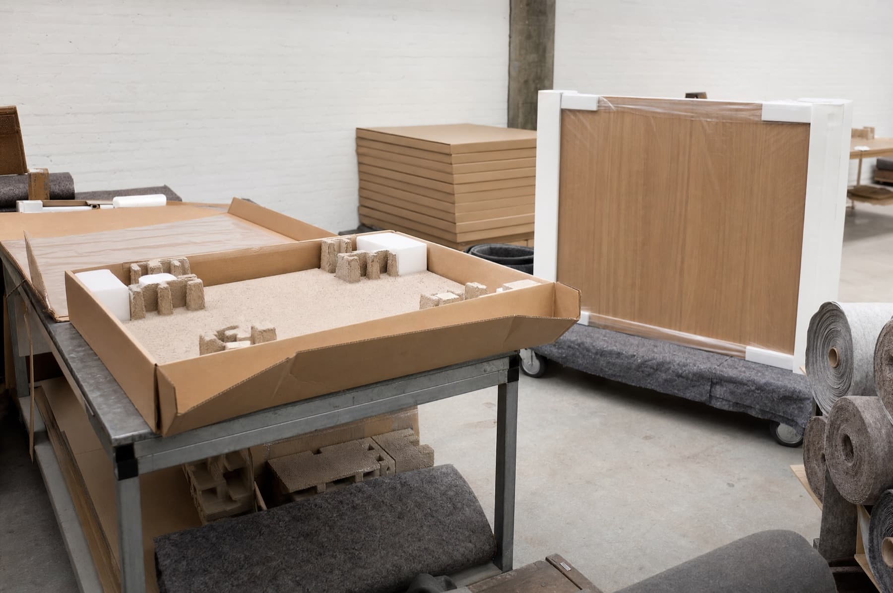 Poste d'emballage : carton plat ouvert sur un établi d'acier, cales de mousse moulée et plateau de table protégé sur un chariot feutré