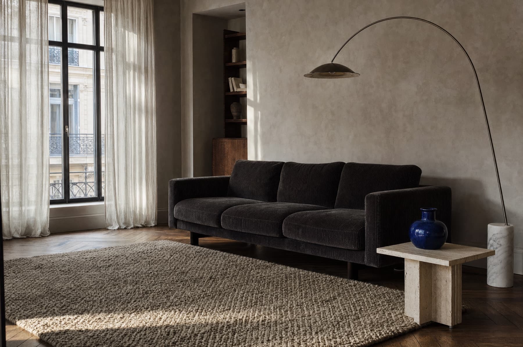 Séjour en fin d'après-midi : canapé en velours côtelé anthracite, tapis de laine, lampadaire à tige arquée et vase en grès bleu sur une table en travertin