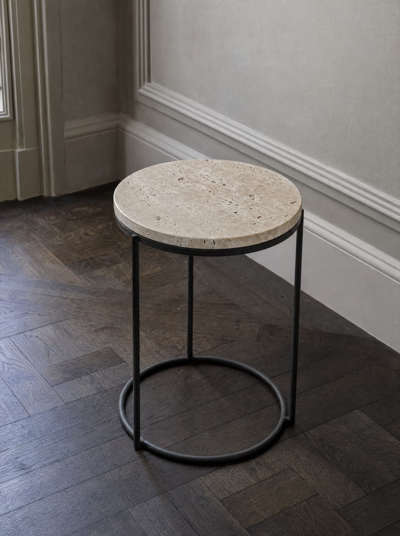 Table d’appoint Sienne, plateau rond en travertin adouci sur piètement tube d’acier noirci, vue de trois quarts sur parquet